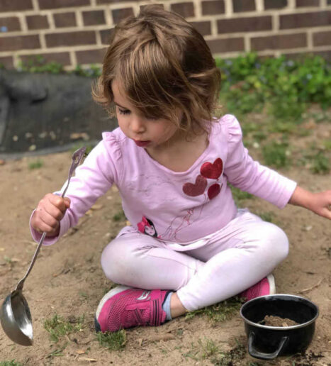 A young girl plays with a ladle and pot in the dirt.