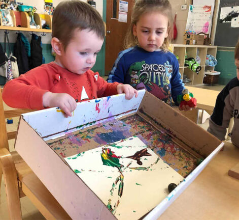 Three children use marbles to paint a picture in a box.