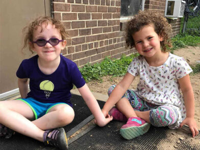 Two young girls sit outside and hold hands.